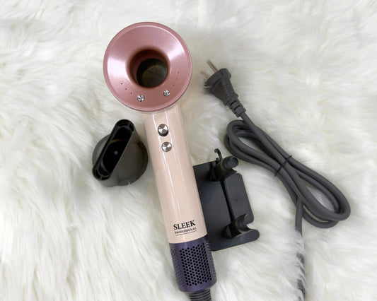 A sleek hair dryer in blush pink, cream, and purple, shown with its black concentrator nozzle, charging dock, and power cord on a white faux fur background.
